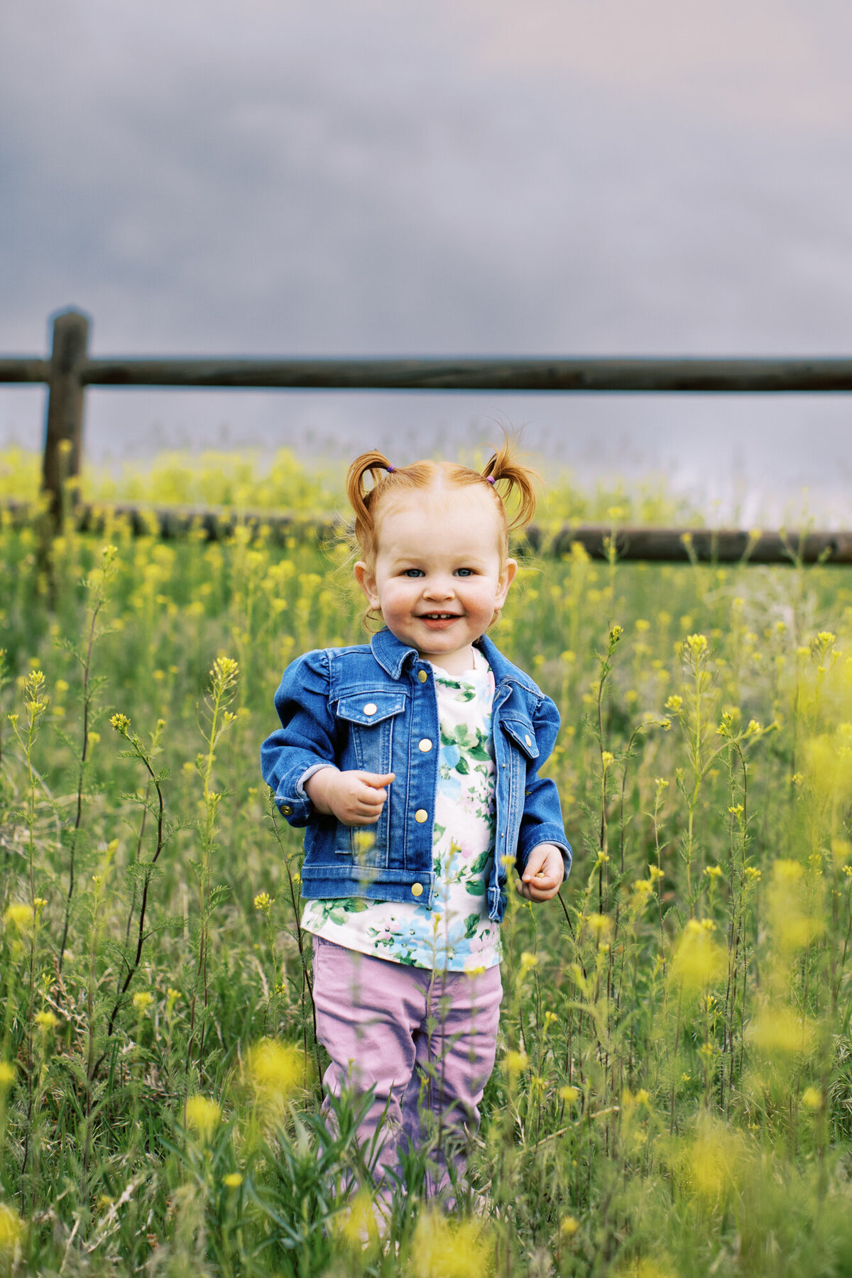 a toddler girl stands in tall green grass with yellow flowers growing