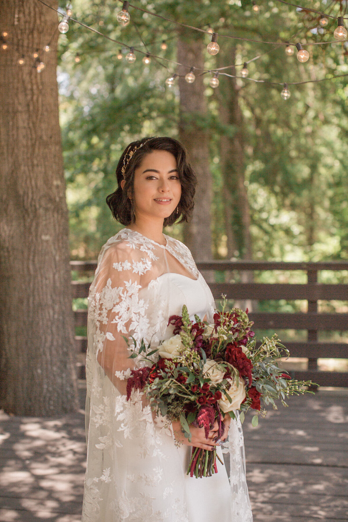 A bride with a gorgeous lacy vail smiles for the camera while holding a stunning bouquet of red flowers