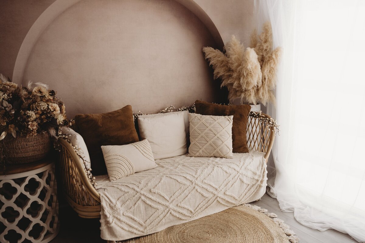 A beige seating area with a cushioned sofa adorned with brown and white pillows, decorative dried plants, and a round woven rug on the floor—perfect for cozy maternity photography sessions.