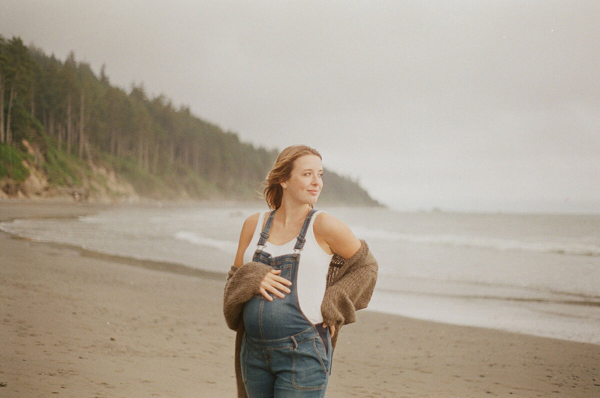 Pregnant mother in denim overalls holding belly in front of PNW coast