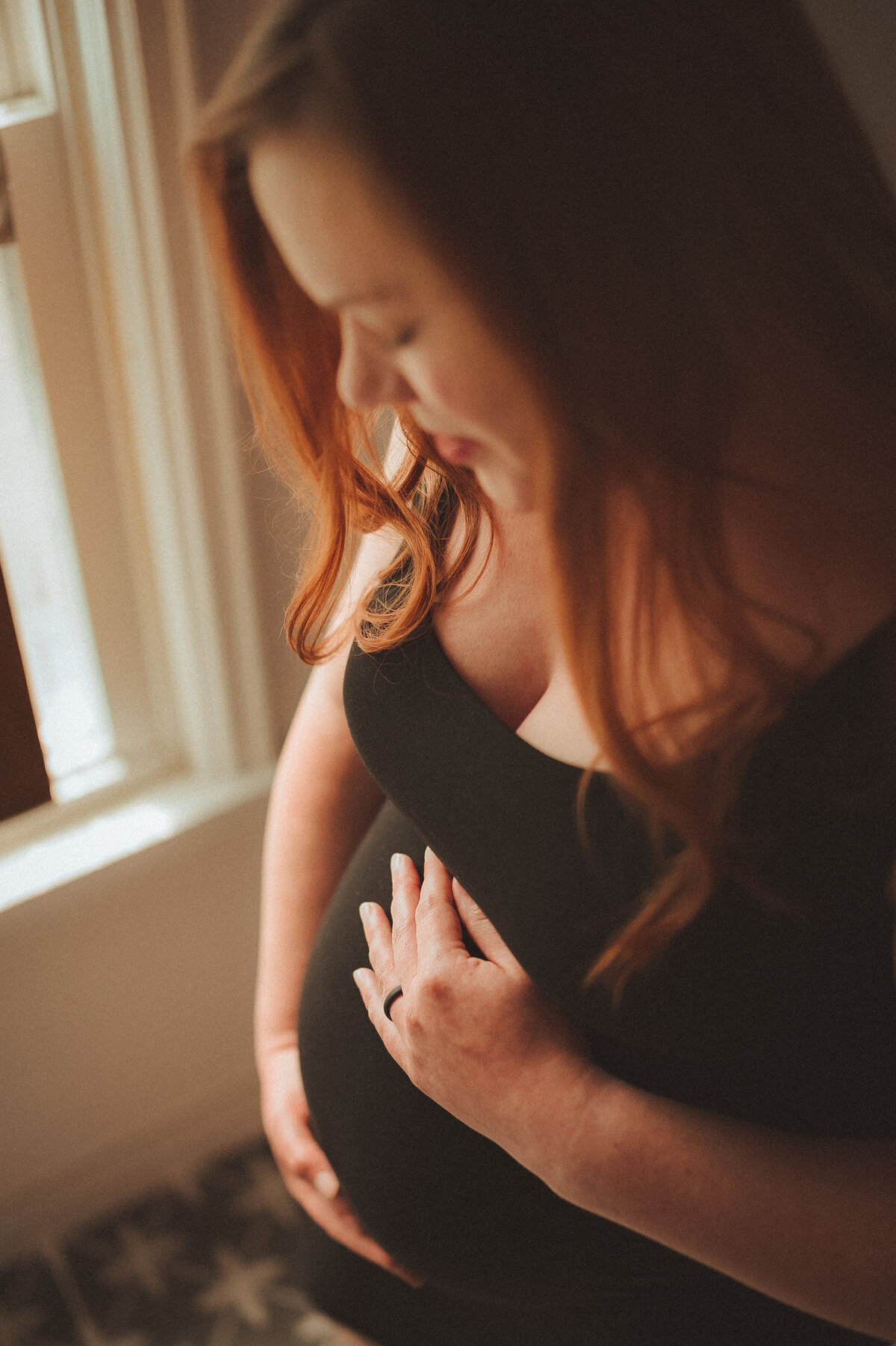 In the heart of Robbinsdale, this intimate maternity portrait captures a mother's deep connection with her growing baby. Bathed in soft, natural light from a nearby window, she gazes lovingly at her pregnant belly, embodying the beauty of this precious moment in her own home.