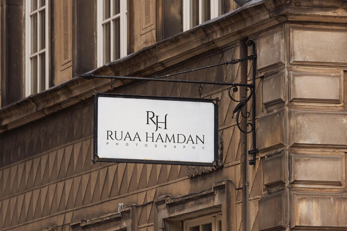 Metal hanging shop sign for “Ruaa Hamdan Photography” mounted on an ornate bracket on a historic stone building facade.