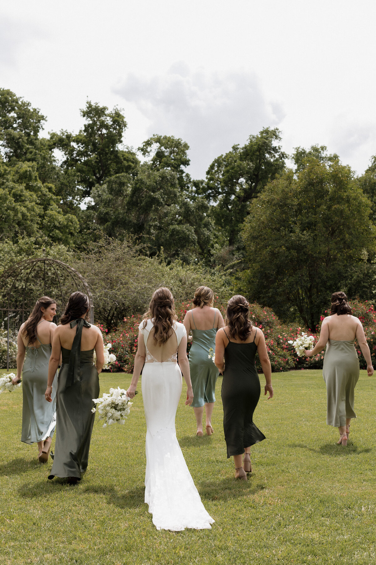 A bride and her bridesmaids walk across a lush green field, dressed in elegant sage and olive gowns, heading toward the wedding ceremony.