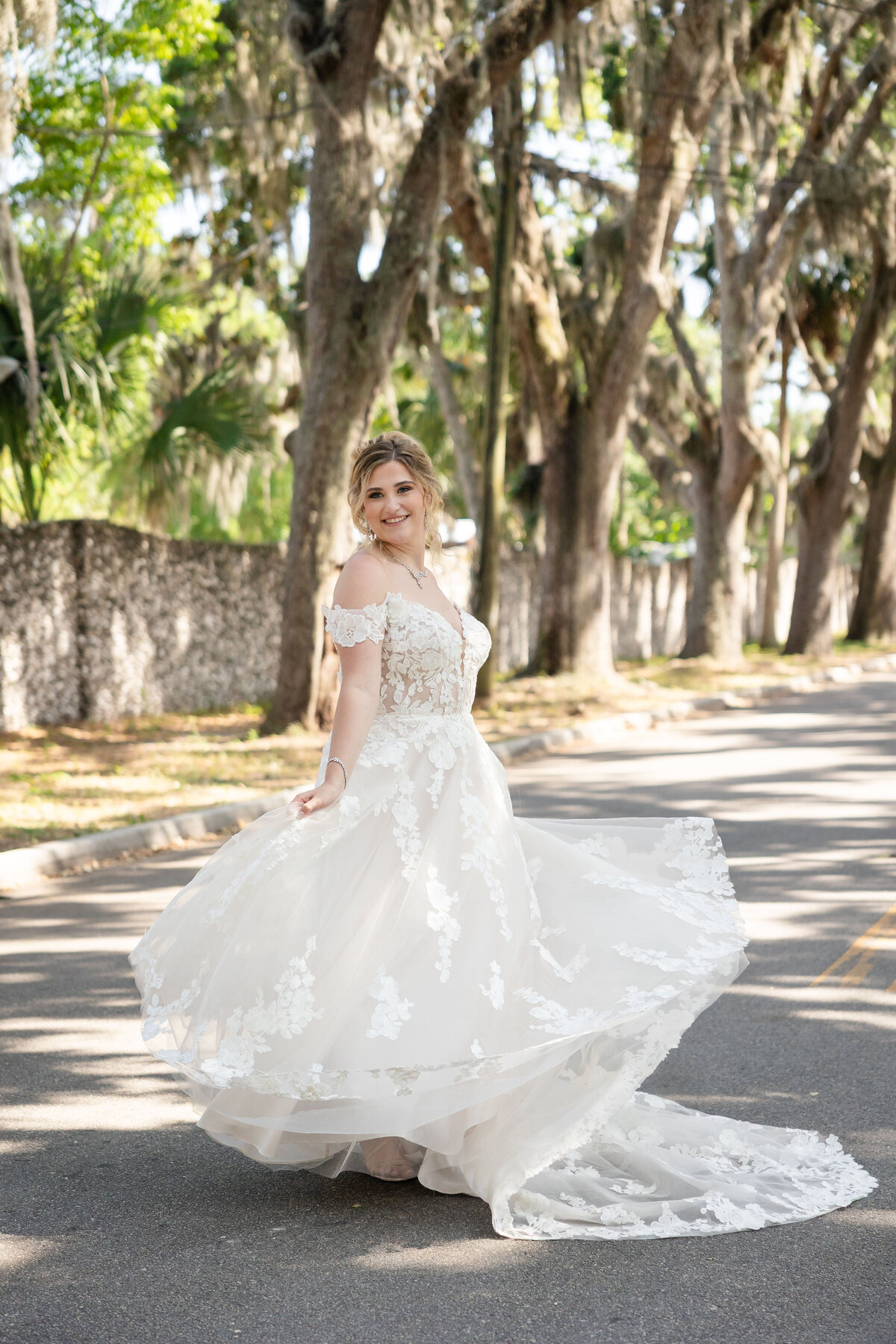 joyful bride twirls in her lace wedding gown on the iconic Magnolia Street in St. Augustine, Florida. Surrounded by moss-draped oak trees and the historic stone walls near the Fountain of Youth, she radiates happiness and grace. Her off-the-shoulder dress flows beautifully in the sunlight as she smiles at the camera. Captured by a wedding photographer who specializes in timeless St. Augustine wedding photography with true-to-color, romantic storytelling.