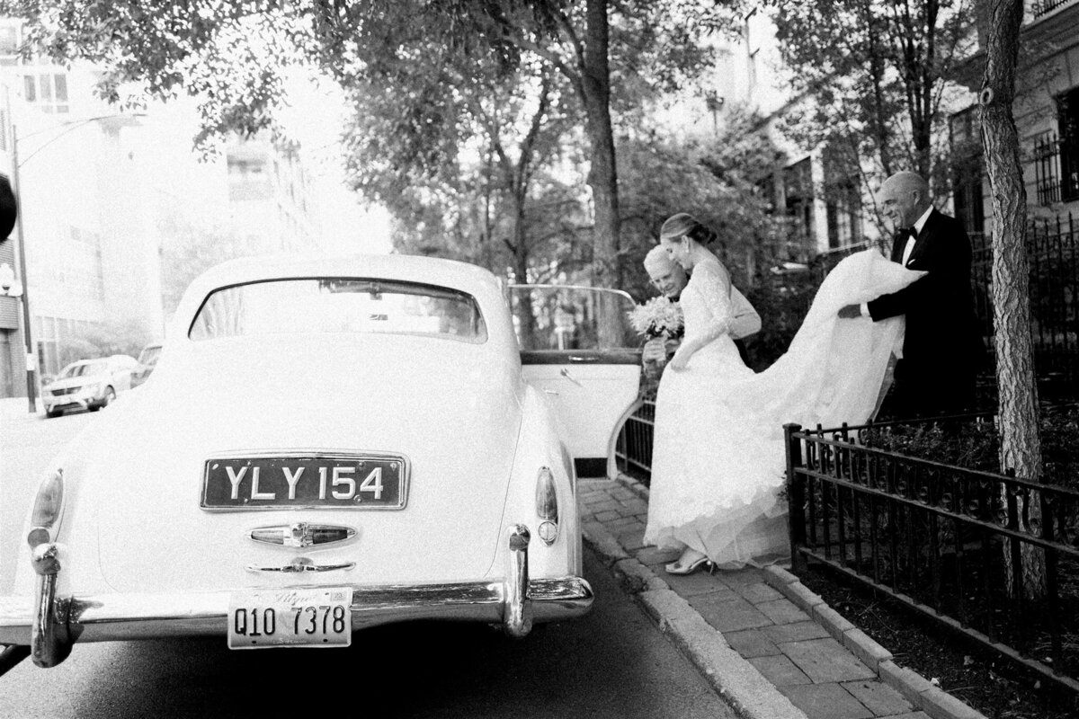 A black and white picture of the bride getting into the old antique car with the help of her mother and father. 
