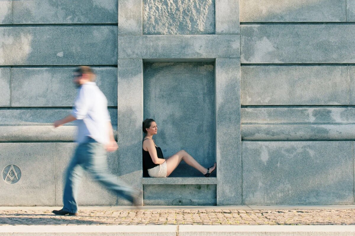 A woman sitting in a recessed niche of a stone wall while a blurred figure walks past