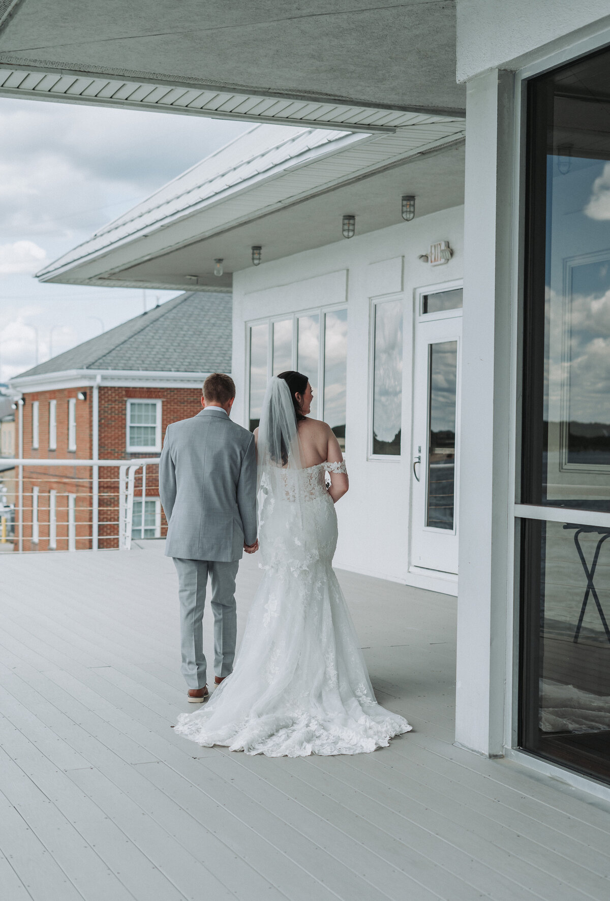 A newlywed couple, with the bride in a long lace gown and the groom in a grey suit, walks hand-in-hand away from the camera on a white pier at the Lesner Inn in Virginia Beach, captured by Quinn Photography LLC.