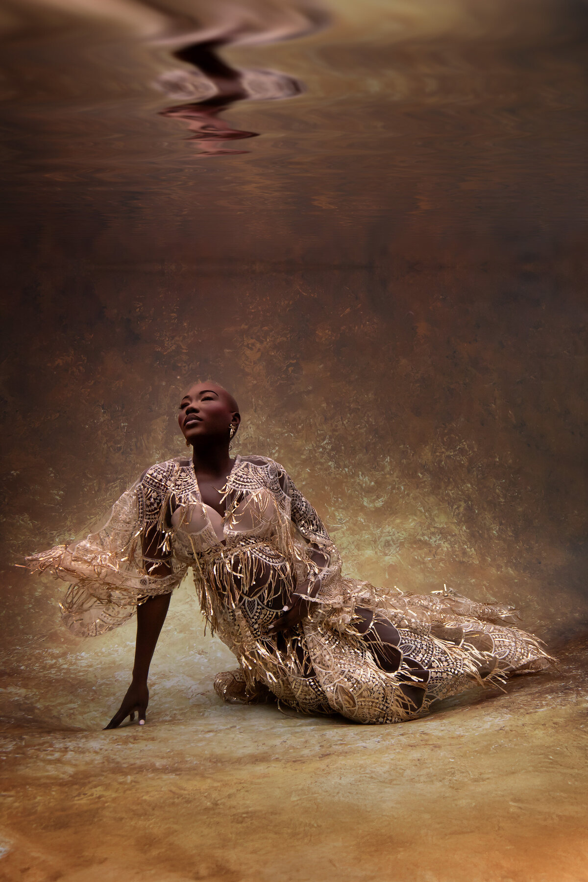 A woman adorned in an intricate gold-fringed, sheer lace gown poses gracefully underwater against a luminous golden backdrop. Her serene expression and confident posture exude elegance and strength, while the delicate embellishments of her outfit shimmer beautifully in the warm, ethereal light.