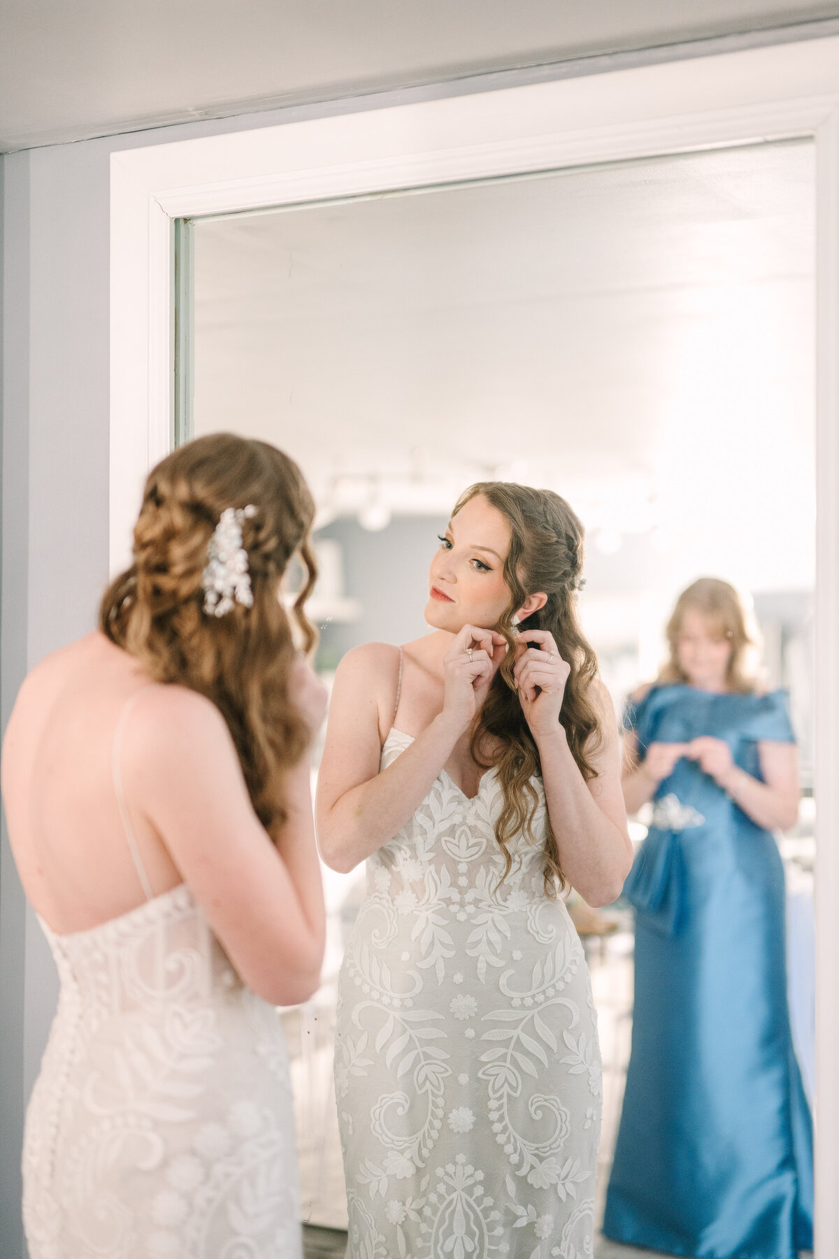 a bride in her wedding gown looking at herself in a full length mirror while the mother of the bride in a blue gown watches from behind her in the Willowbrook wedding venue preparation suite