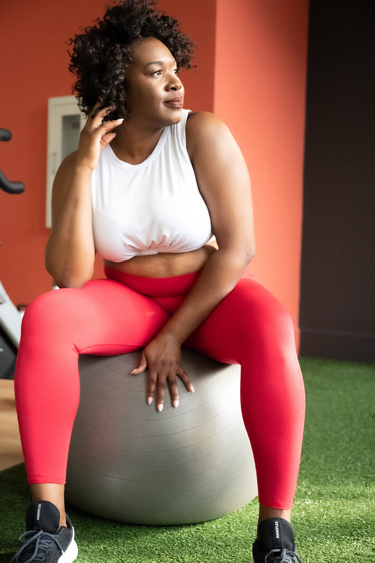 A portrait of a woman in a gym setting, setting on a big grey ball looking a the window