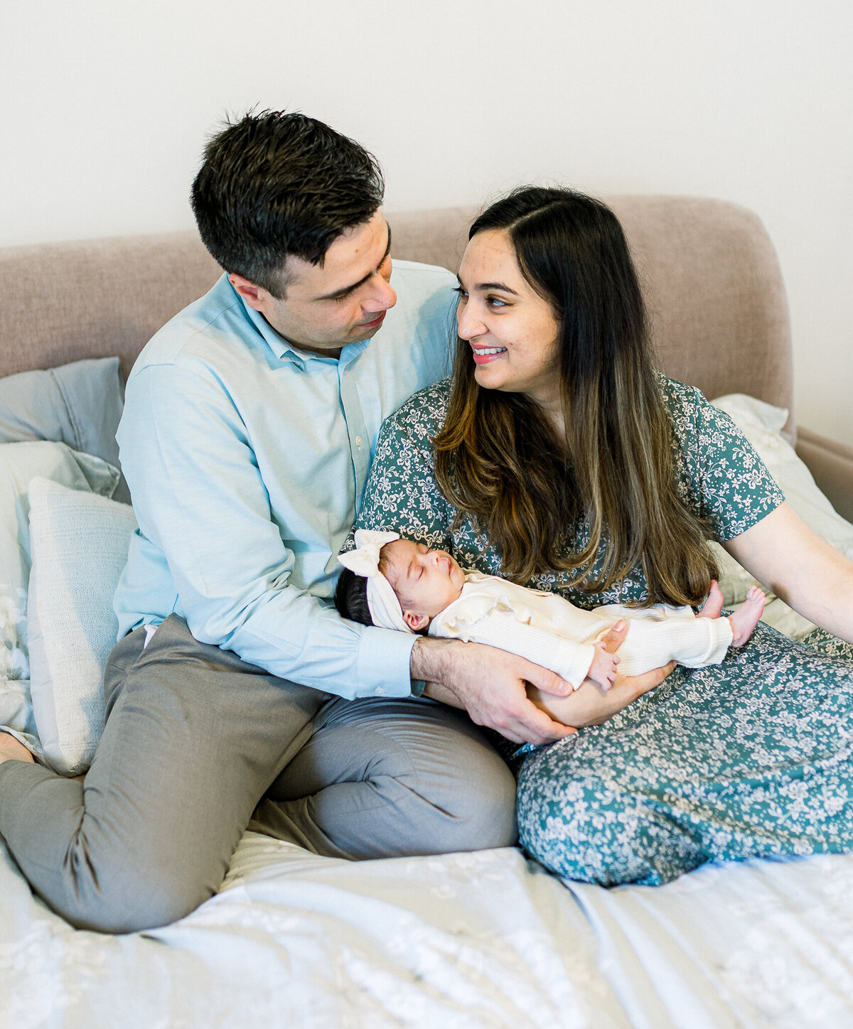 New parents sit on their bed, gazing at the camera while cradling their sleeping newborn daughter dressed in an ivory outfit with a soft bow headband. A cozy in-home newborn photography session capturing love and family connections.