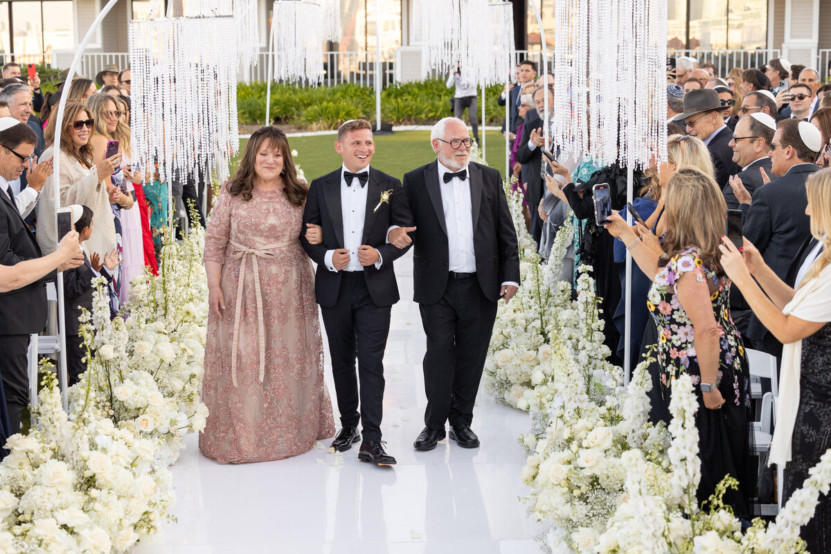 A groom walking down an aisle with his parents