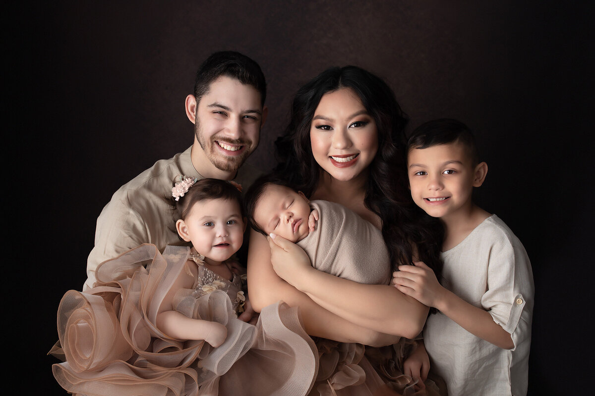 Luxury family portrait in a fine art photography studio. A radiant mother cradles her sleeping newborn, surrounded by her smiling husband and two older children in coordinating neutral tones. The little girl wears an elegant tulle gown with floral embellishments, adding to the timeless and sophisticated aesthetic. Captured in a high-end family and newborn photography session serving Dallas and surrounding areas.