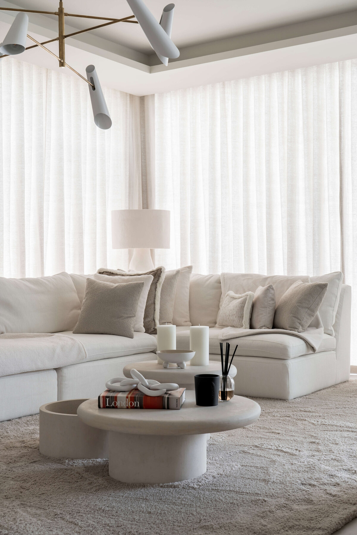 A spacious, modern living area. A large white couch with neutral throw pillows sits on a soft, textured cream rug. Behind the couch are floor-to-celiing cream drapes extending the length of the wall.