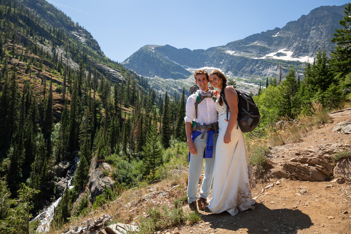 A bride and groom stand smiling at the camera on a dirt trail in Montana on their adventure elopement day.