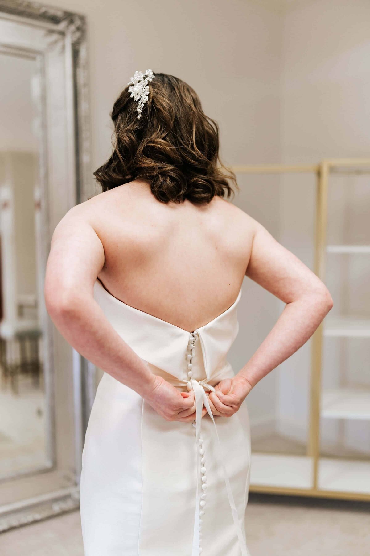 A woman in a strapless white dress ties the back of her gown. Her hair is styled in loose waves with a jeweled hairpiece. She stands in a bridal suite with a large mirror and empty shelves in the background.