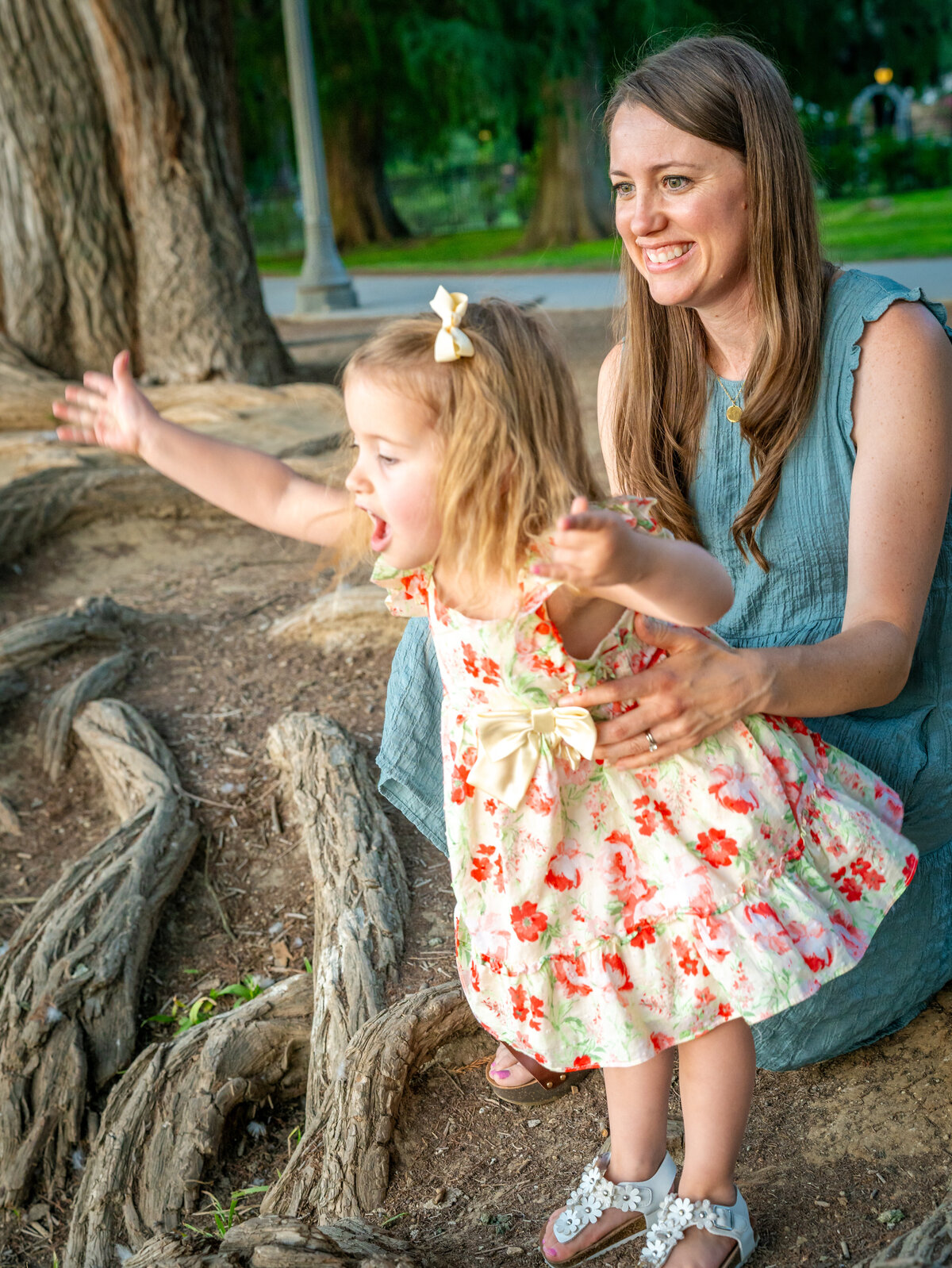 A smiling mother in a sea-foam green dress, holds her exuberant toddler daughter around the waist as the little girl in the flowered sundress leans out over a park drop-off with her arms spread white and her mouth gaping open. Photo by SAVI Photography - Photographer in Riverside