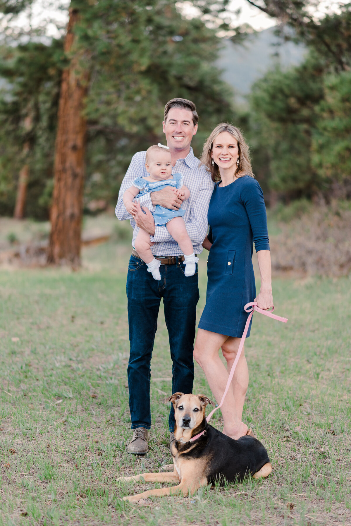 family outdoor photos with father holding a small infant while the mother holds their dog as they pose together in the woods with denver family photographers