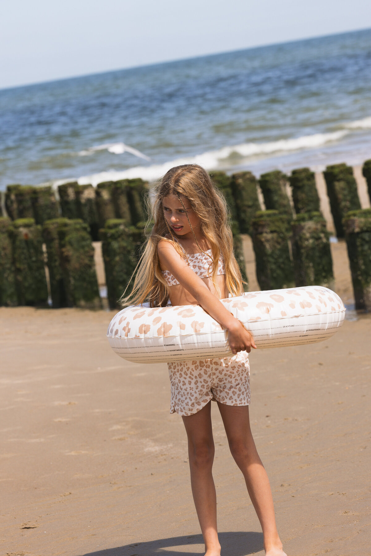 Young girl standing on the beach holding a white and beige leopard print Swim Essentials float, wearing a matching bikini set, with the sea and moss-covered wooden poles in the background.