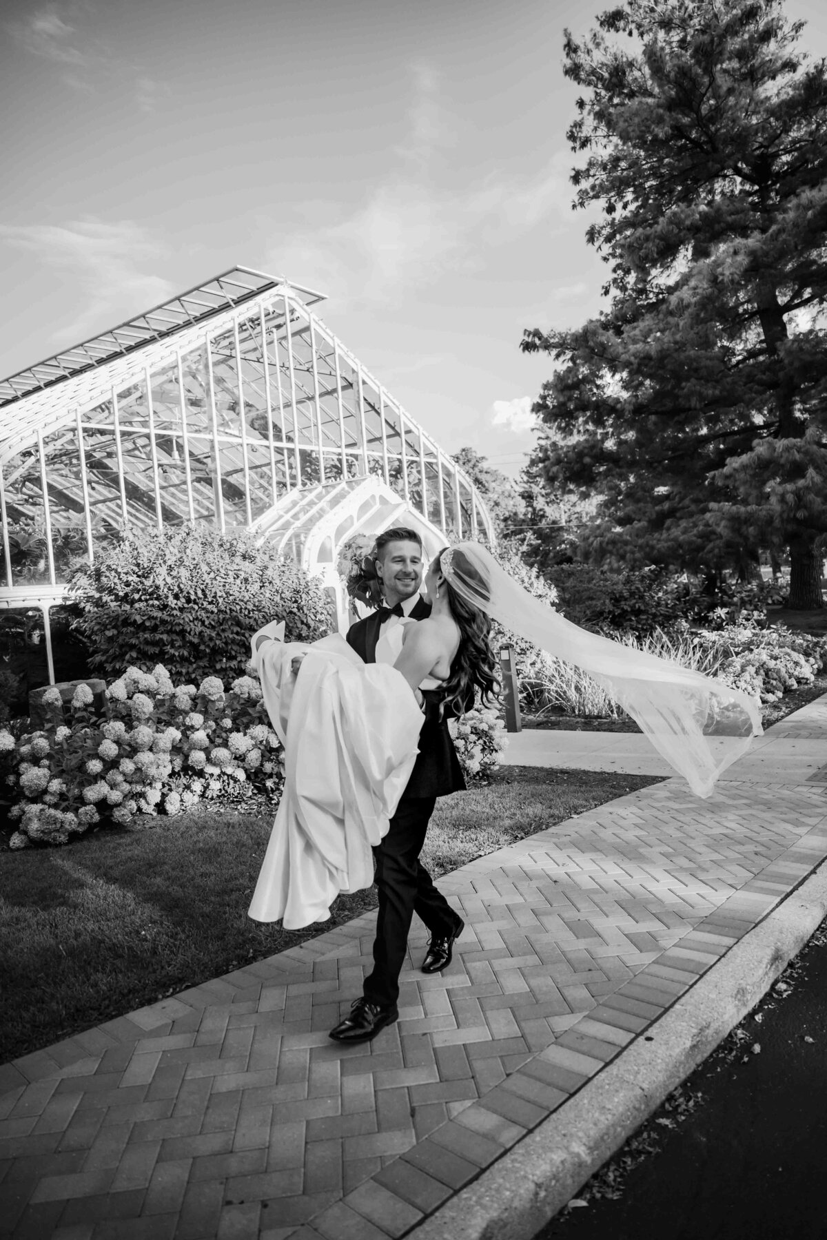 Groom carries bride in front of greenhouse