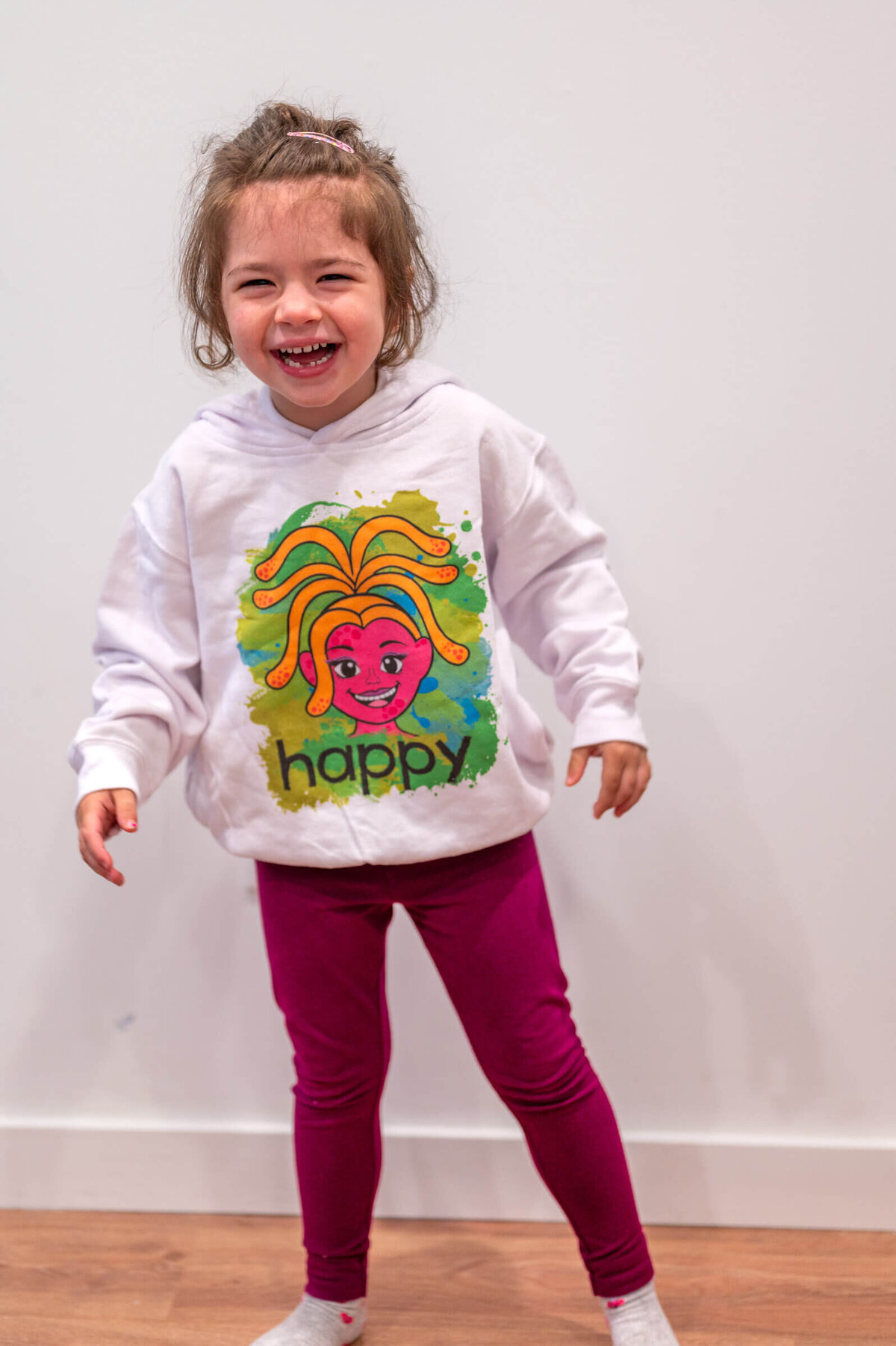 Girl laughing while wearing a white sweatshirt with a colorful Luua Play branded character and "Happy" on it.