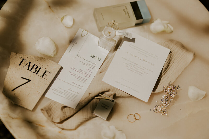 Ensemble de papeterie de mariage entouré de bijoux, alliances, pétales de fleurs. Ambiance minimaliste beige et blanche. Scène imaginée par laura Termeau, photographe professionnelle en Vendée.