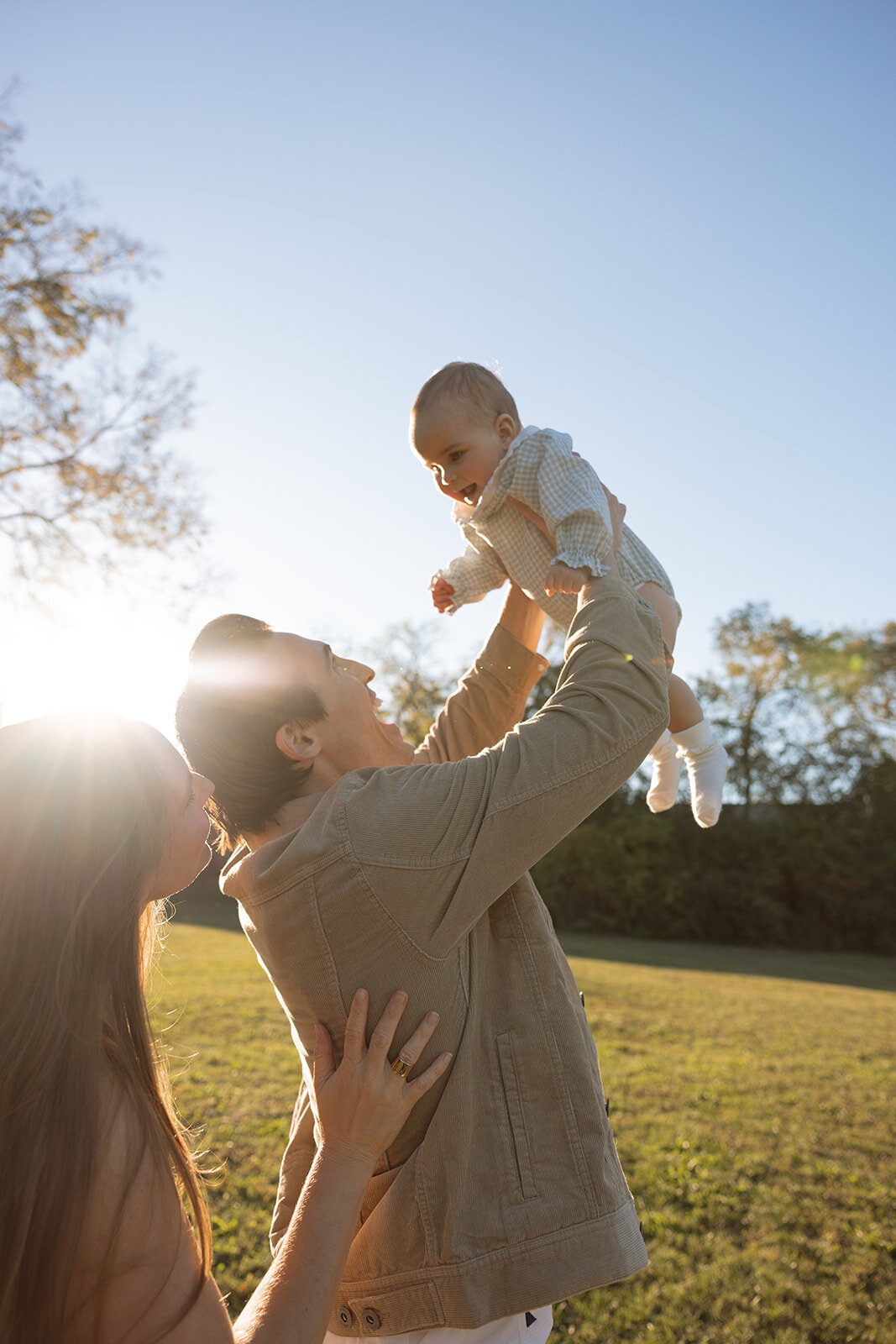 Dad joyously lifts infant high in the sky while infant smiles and sunshine douses them in the golden hour.