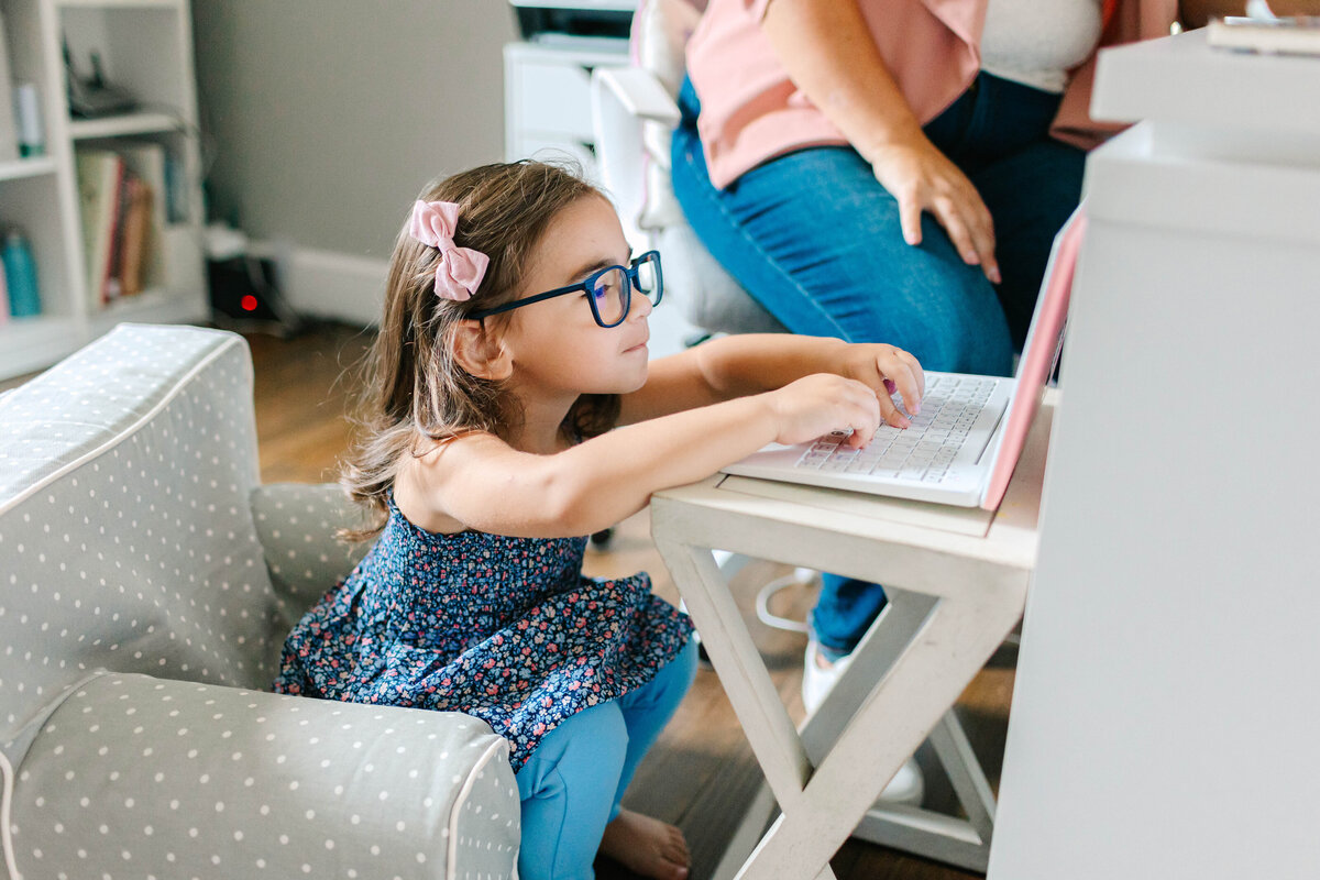 A young girl with glasses and a pink bow in her hair types on a pink laptop while sitting on a small polka-dotted chair. An adult, partially visible in the background, sits nearby at a desk.