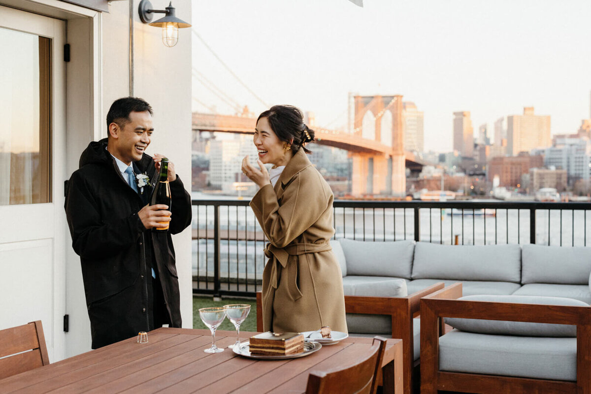 The bride and the groom are laughing together, with a slice of cake and a bottle of wine. The Brooklyn Bridge is in the background.