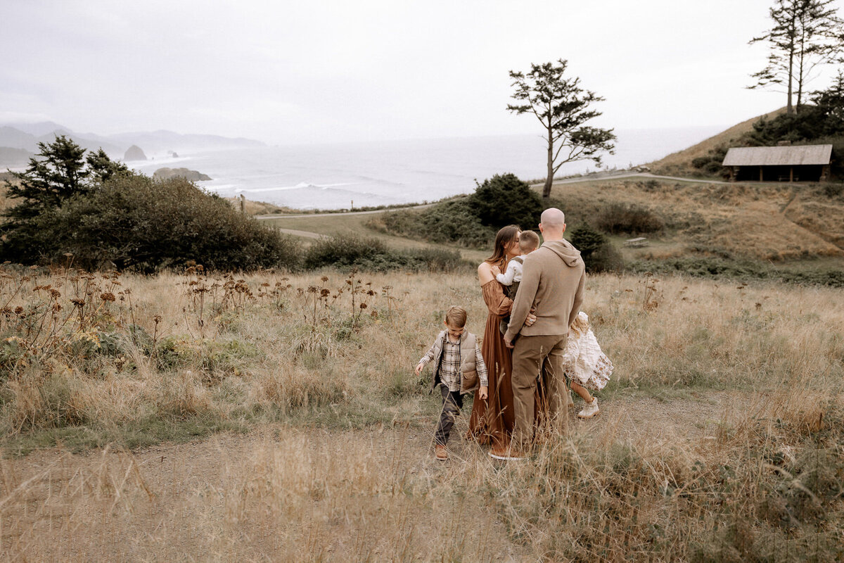 "A beautifully candid family moment captured in nature, with parents and their children walking through a golden field overlooking the ocean. The family's earthy-toned outfits blend seamlessly with the serene coastal backdrop, creating a timeless and heartfelt scene. Photographed by Mary Wiebe Photography, Lubbock family photographer