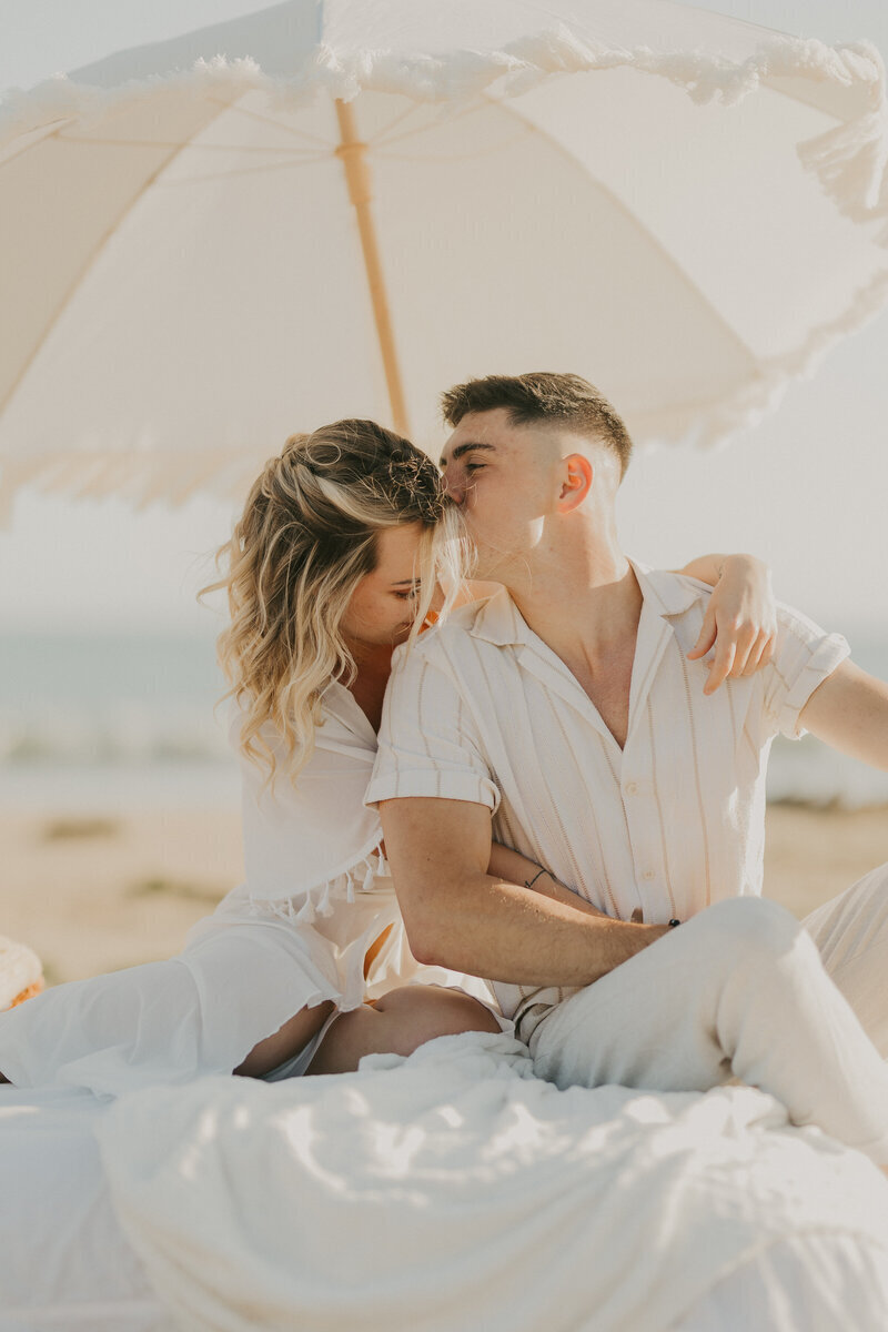 Couple assis sur un matelas recouvert d'un drap blanc, s'enlaçant sous une ombrelle  à la plage pendant une séance photo à la plage en vendée.