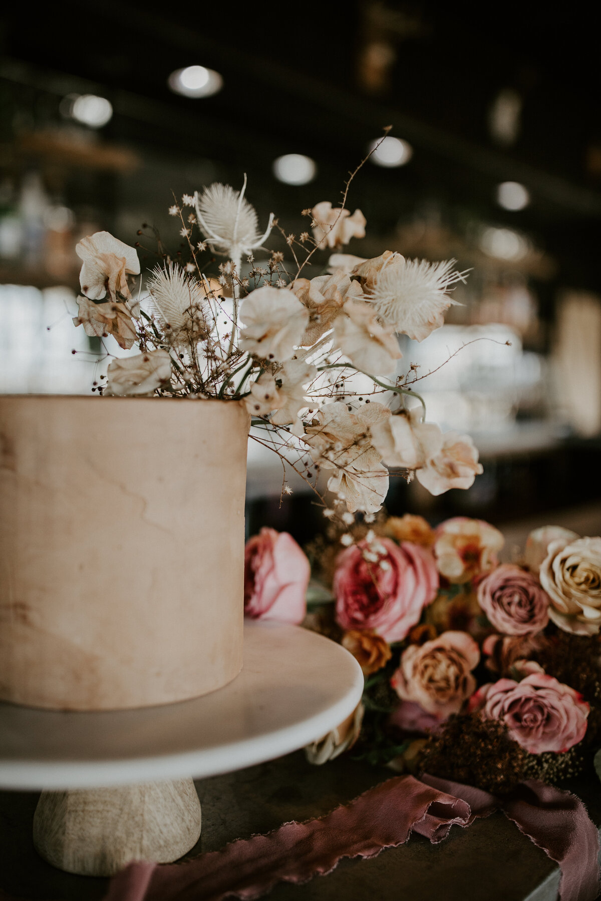 A cream-colored cake with ivory flowers sitting on a marble cake stand with pink and mauve flowers.