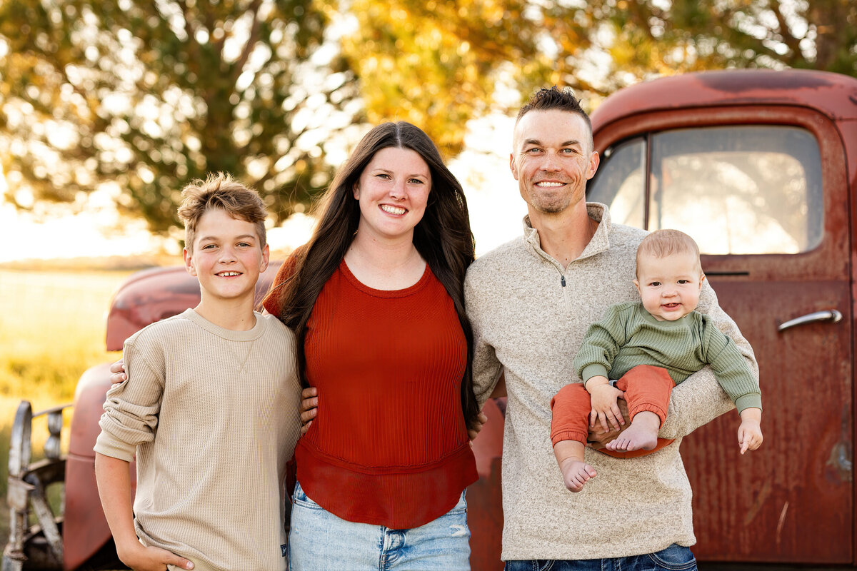 Mom and dad and their two sons smile at the camera standing in front of a vintage red farm truck.