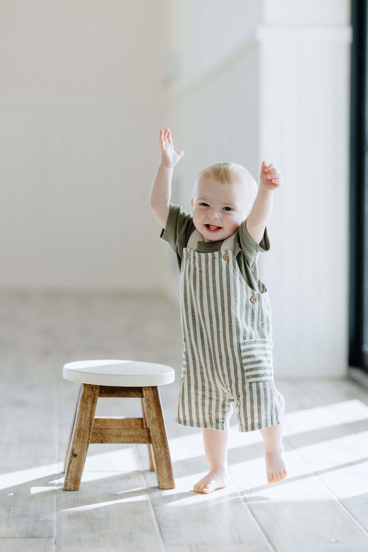 One year old son in stripped overalls and green shirt standing by wooden stool