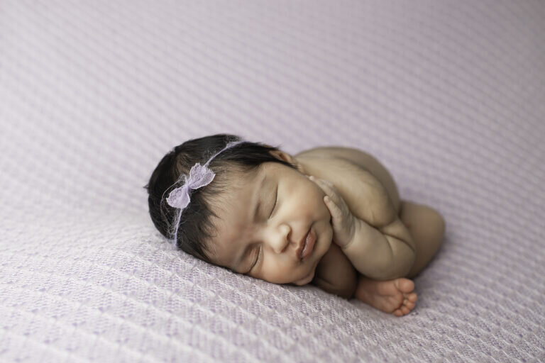 Newborn baby girl with a full head of hair, curled up in a sweet, folded position, peacefully resting in a soft and cozy setup