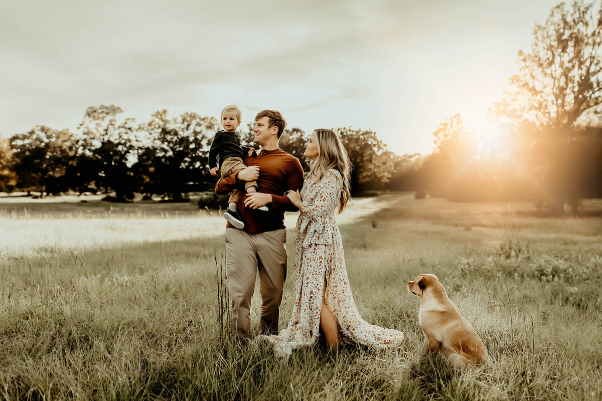 A family of three and a dog stand in a grassy field during sunset. The father holds a child, while the mother stands beside them. The dog sits nearby, looking into the distance.