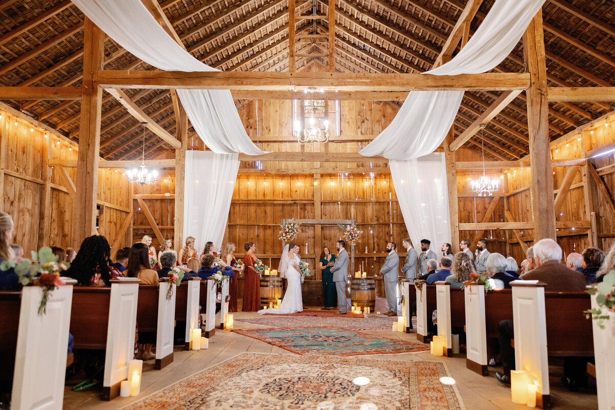 Wedding ceremony in rustic barn with draped white fabric, chandelier, and floral arch at The Hummingbird Chateau.