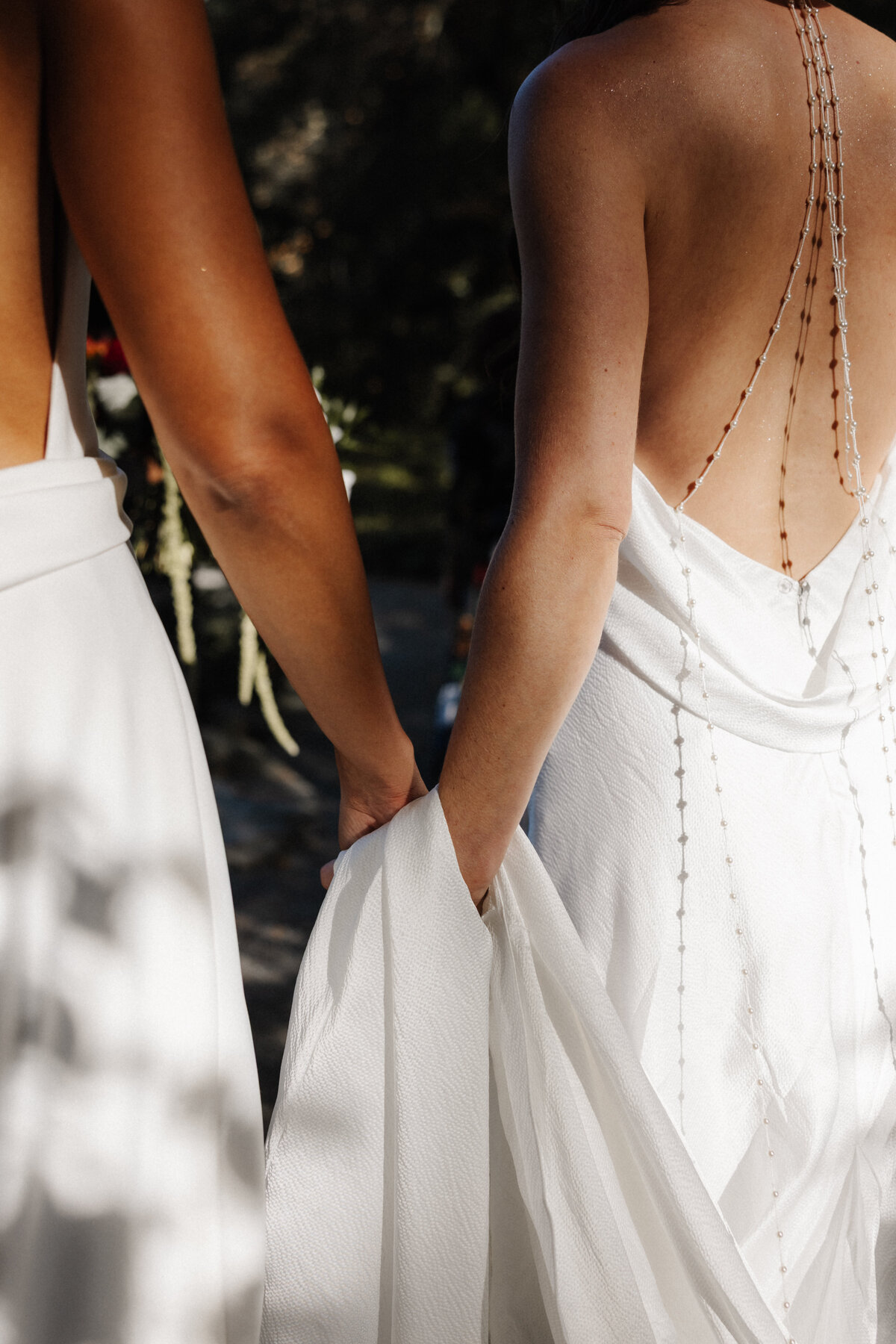 Close-up of two brides holding hands, showing the backs of their wedding dresses and pearl straps.
