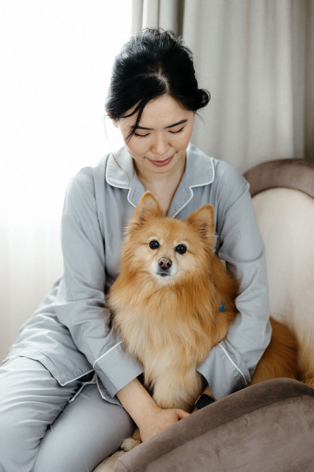 A woman in her pajamas is sitting while hugging and looking down on her brown dog.