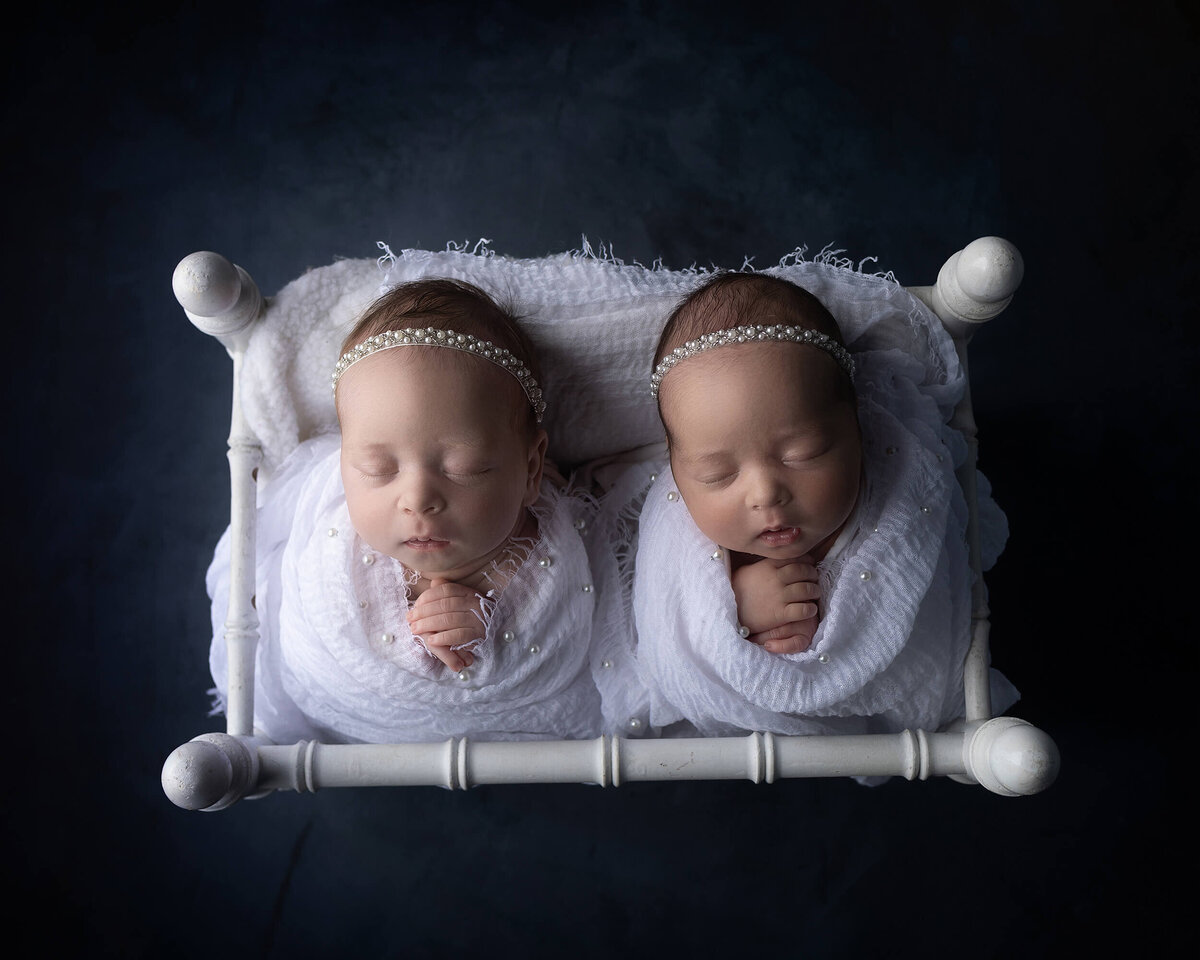 Sleeping twin newborn girls wearing all white and pearls, in white bed on navy blue background