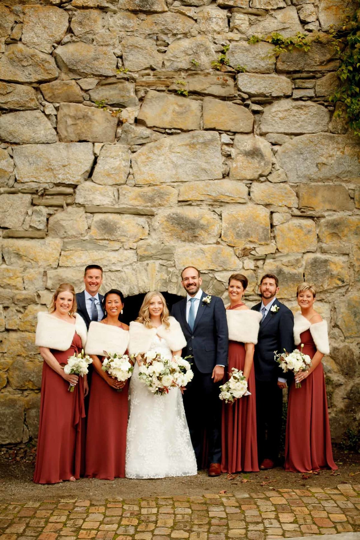 wedding party with brides maids wearing rust colored dresses with white cape, holding white flower bouquets