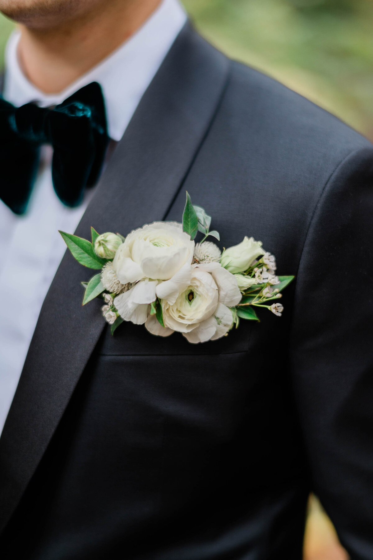 A detailed shot of the groom’s boutonnière, a cluster of white ranunculus and greenery pinned to the lapel of his black tuxedo
