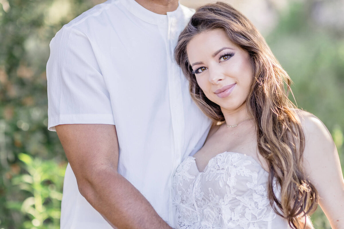 A woman in a white lace dress stands next to a man in a white shirt. The woman is smiling subtly, with long hair and soft makeup. They are outdoors with greenery blurred in the background.