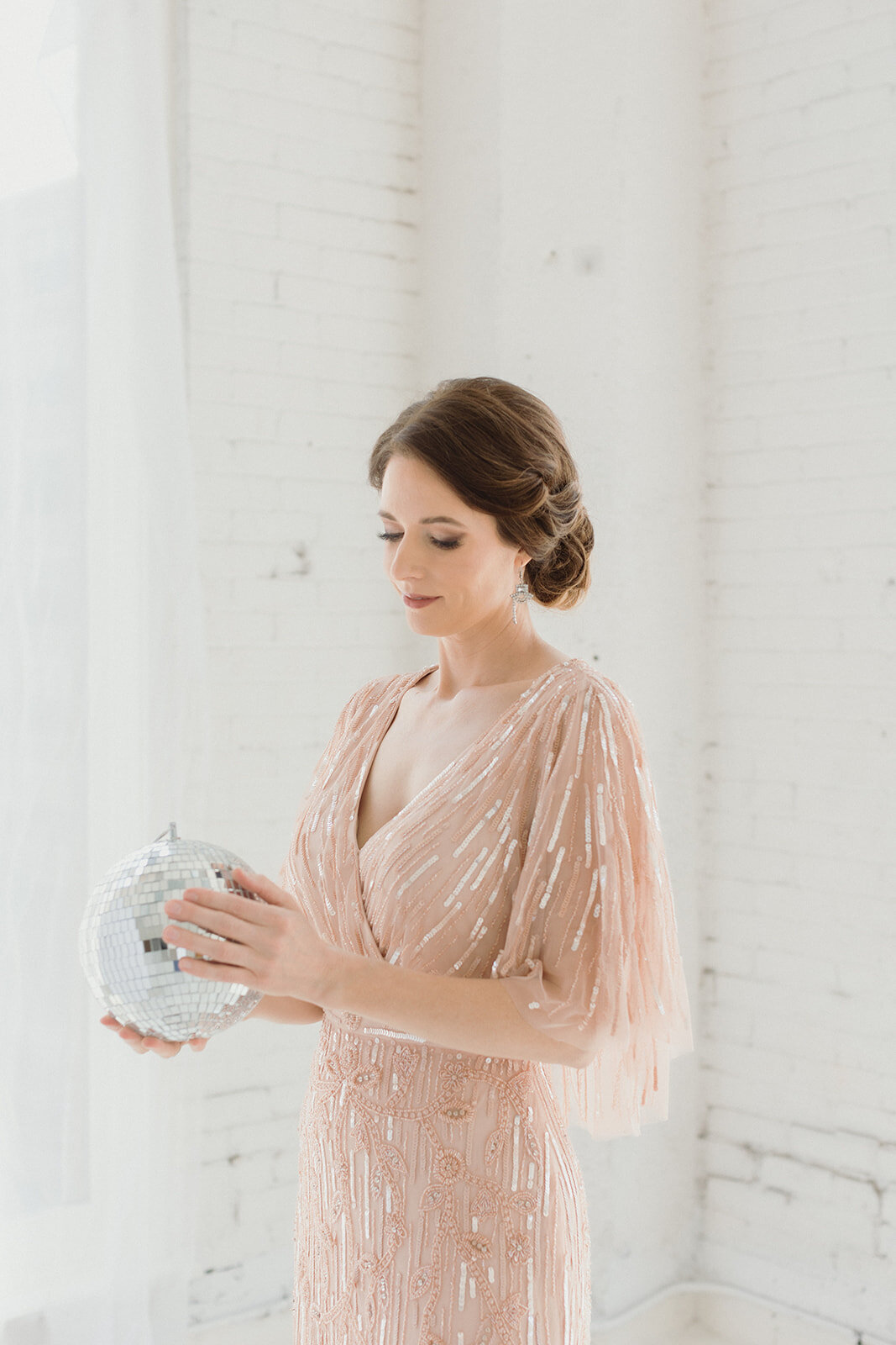 Woman wearing light pink sparkly gown, holding a disco ball and looking at it.