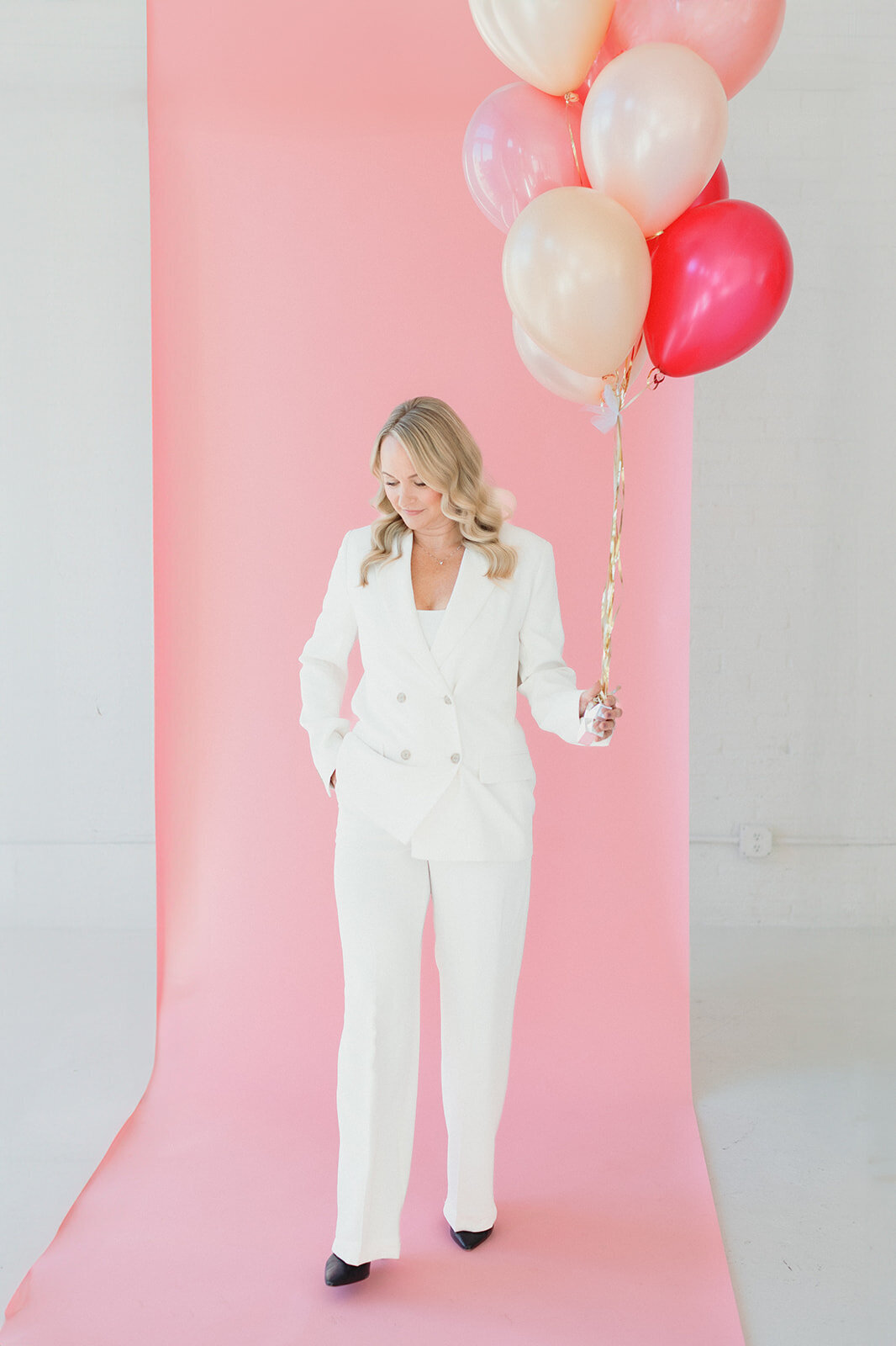 blonde woman wearing white suit is walking in front of pink backdrop and carrying large pink and white balloons. She is looking down toward the ground.