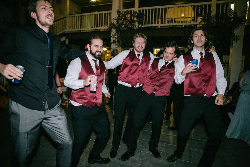 Five men in dress shirts and vests stand close together with arms around each other, smiling and enjoying themselves.
