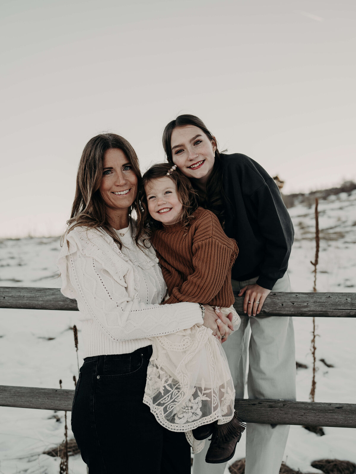 A portrait of a mother with her two daughters during an outdoor winter photography session. All three girls were coordinating color, pallets of Browns and creams as they smile at the camera.
