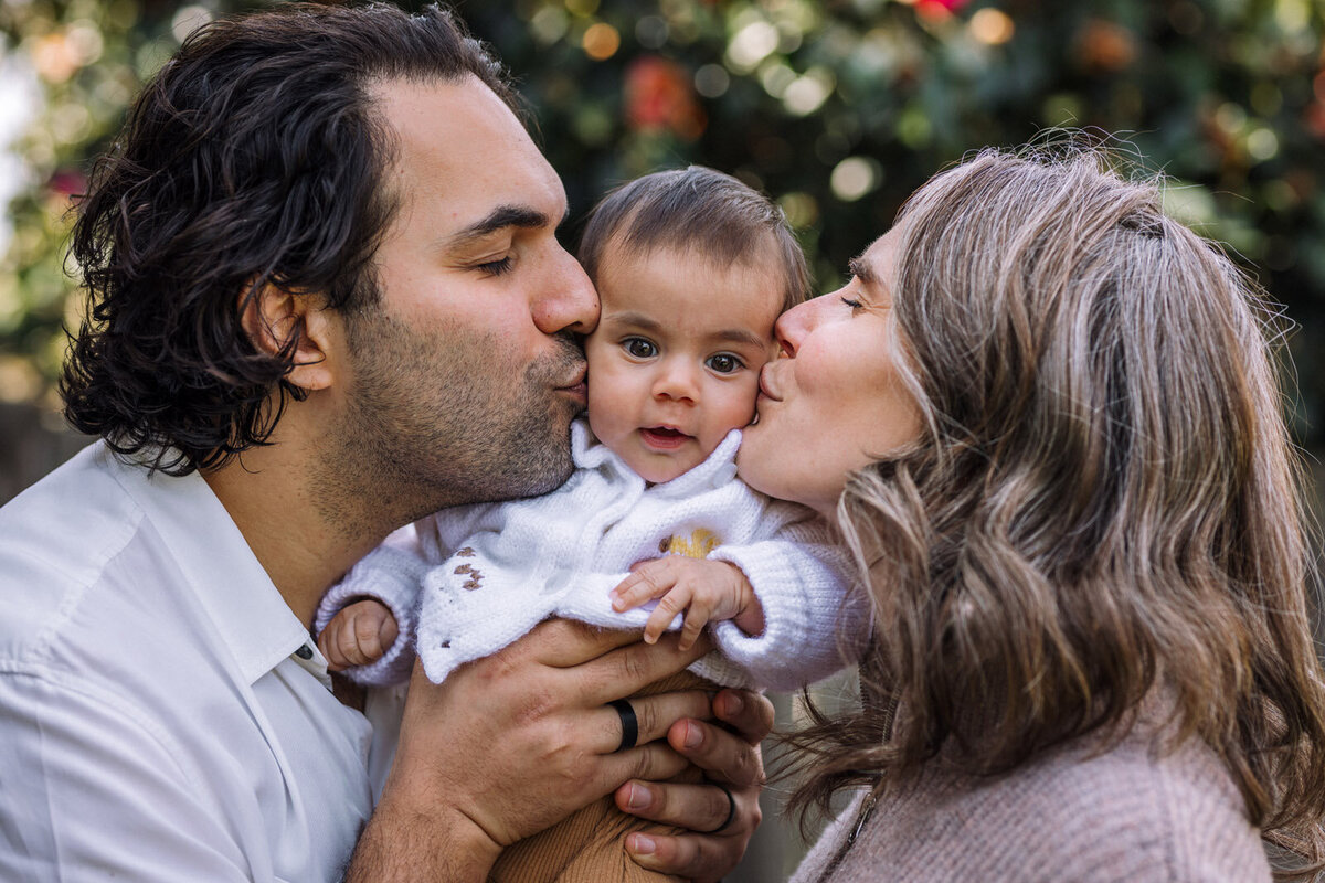 mom and dad holding baby together and kissing her cheeks in Alameda backyard