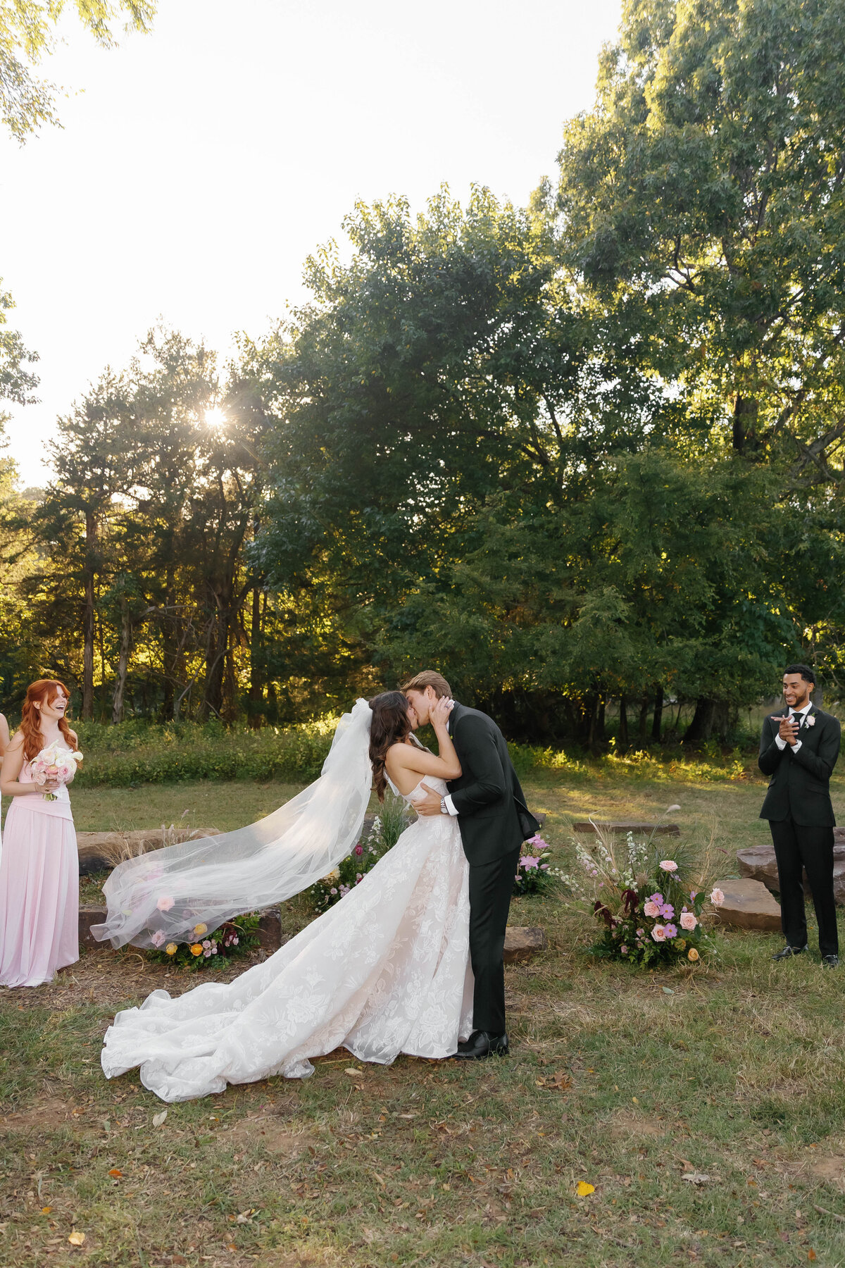 Bride and groom share their first kiss as newlyweds in an outdoor forest-edge ceremony, framed by whimsical meadow-style floral arrangements created by an Arkansas wedding florist.