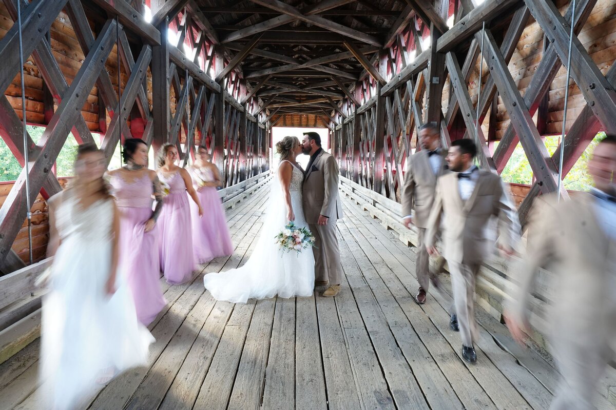 A couple shares a tender kiss on a bridge while their bridal party walks by in the background. The scene captures a joyful and celebratory moment with the couple in the foreground and their friends adding to the festive atmosphere.